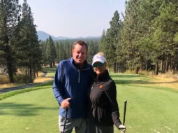 Devin Doyle and his wife pose with their golf clubs on a scenic, tree-lined golf course